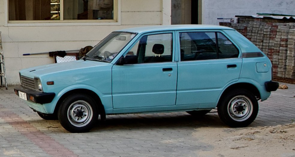 Classic Maruti 800 hatchback parked outside a building, showcasing its simple design and iconic light blue color