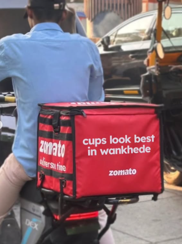 Zomato delivery rider on a bike with a red Zomato delivery bag featuring the slogan “cups look best in Wankhede,” promoting cricket-themed food delivery in Mumbai.