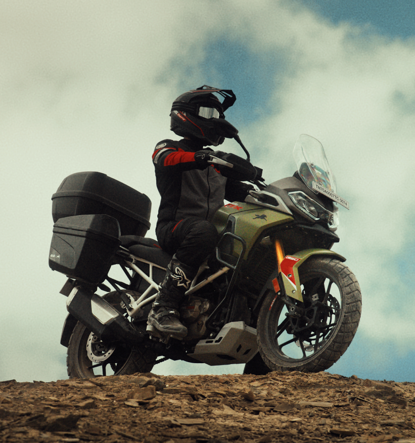 A motorcyclist riding the TVS Apache RTX 300 adventure bike on a rocky trail, equipped with touring boxes, highlighting its off-road strength and adventure-ready design.