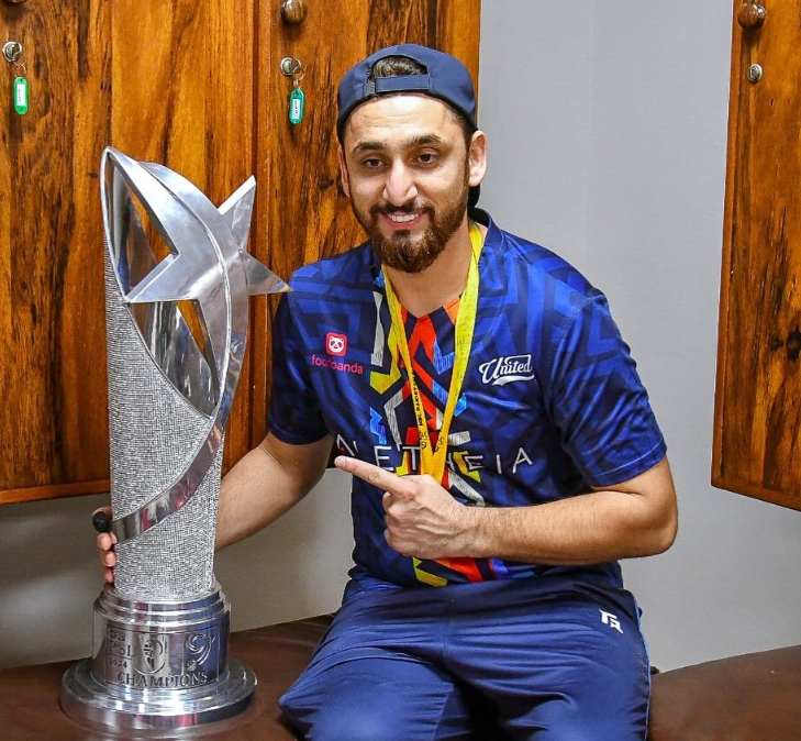 Pakistani cricketer sitting in the dressing room proudly posing with the PSL championship trophy, wearing a blue team jersey and medal around his neck after a tournament victory.