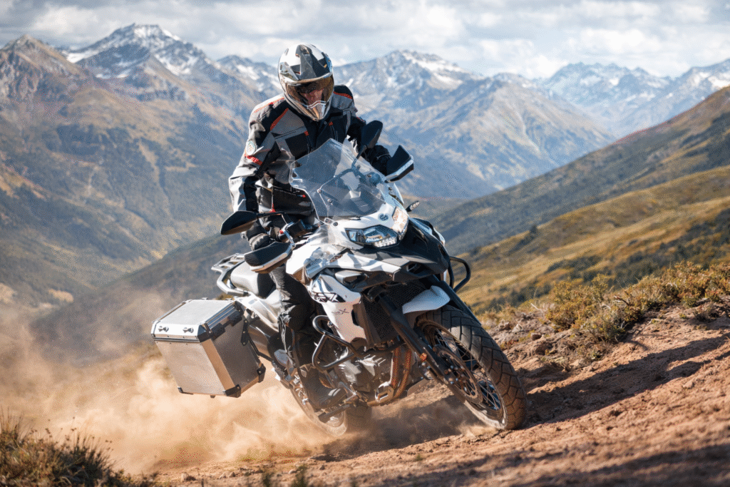 Rider riding a Benelli TRK 502 adventure motorcycle on a mountain trail with hills and snow-capped peaks in the background